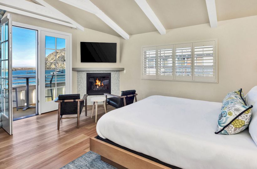 A cozy bedroom features a bed, two chairs, a fireplace, TV, and a balcony with ocean views, all under a vaulted ceiling with white beams.