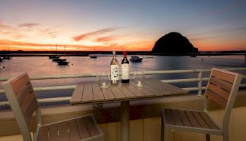 A serene sunset over water with boats, featuring a patio table set with two chairs, two wine bottles, and two glasses, creating a cozy ambiance.