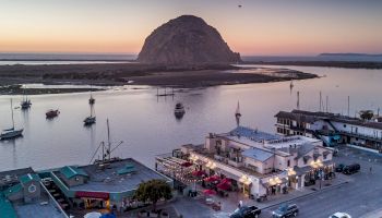 A coastal town at sunset with boats docked in the water and a large rock formation in the distance, featuring waterfront buildings and parked cars.