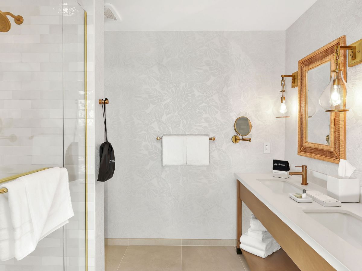 A well-lit, modern bathroom with a glass shower, two sinks, golden fixtures, and matching mirrors above the counter.