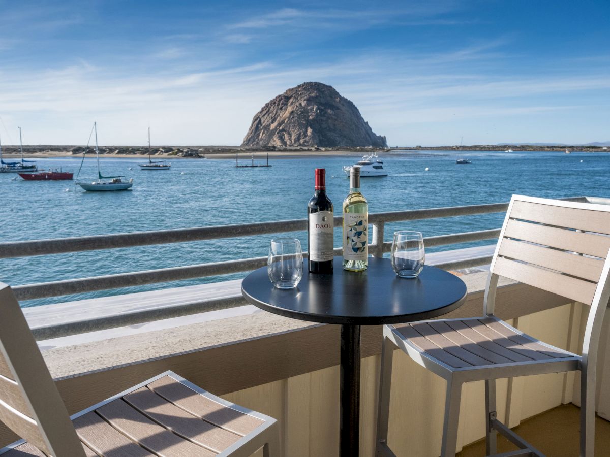 A serene coastal view featuring a table with two wine bottles, empty glasses, and chairs overlooking boats on the ocean with Morro Rock.