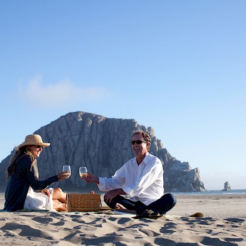 Two people enjoying a beach picnic, toasting with wine glasses, with a large rock formation in the background and clear blue skies.