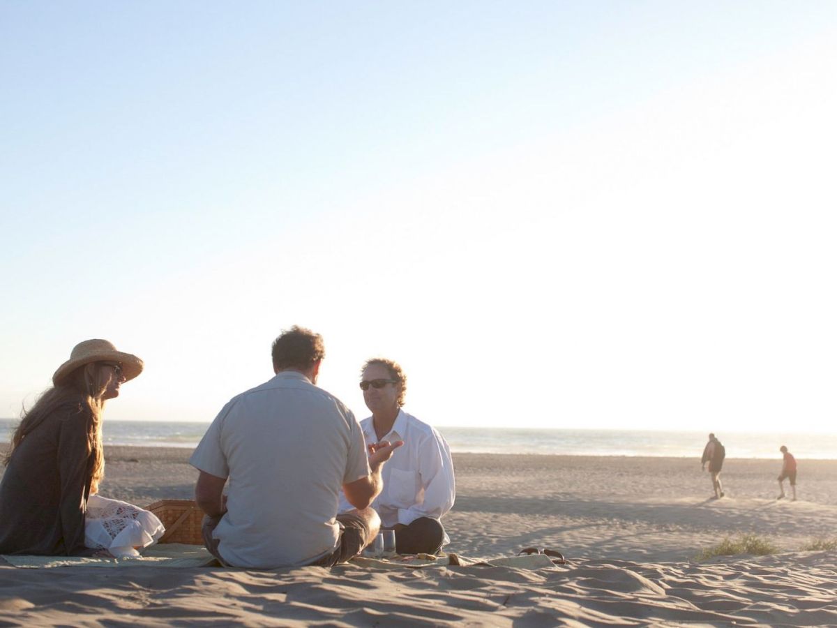 Three people are sitting on the beach, talking while others walk in the distance. The sun is setting, casting a warm glow over the scene.