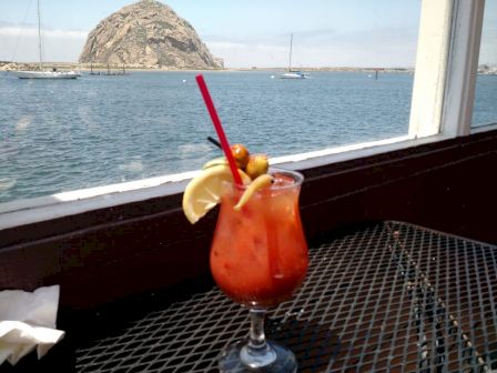 A Bloody Mary garnished with olives and lemon sits on a metal table by a window, overlooking a scenic view of the ocean and a large rock formation.