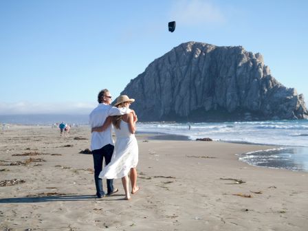 A couple walks on a beach with a large rock formation in the background, while a few people can be seen in the distance near the shoreline.